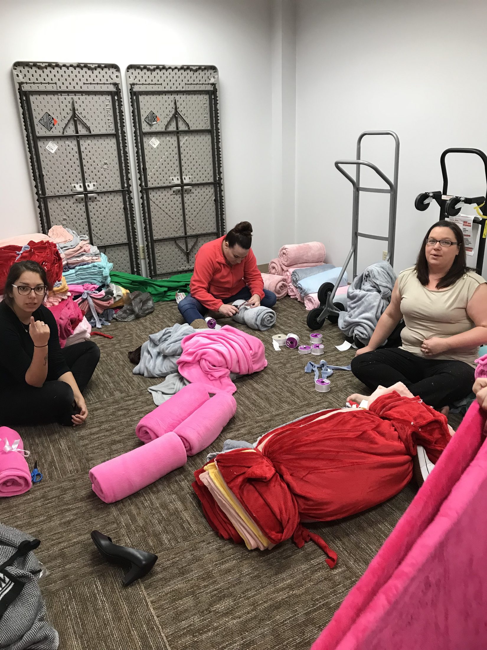 Three volunteers sitting on a carpeted floor folding and rolling colorful fleece blankets for foster children.