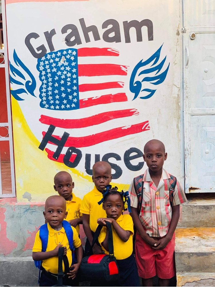 Five children standing in front of a hand-painted Graham House mural with an American flag and blue wings.