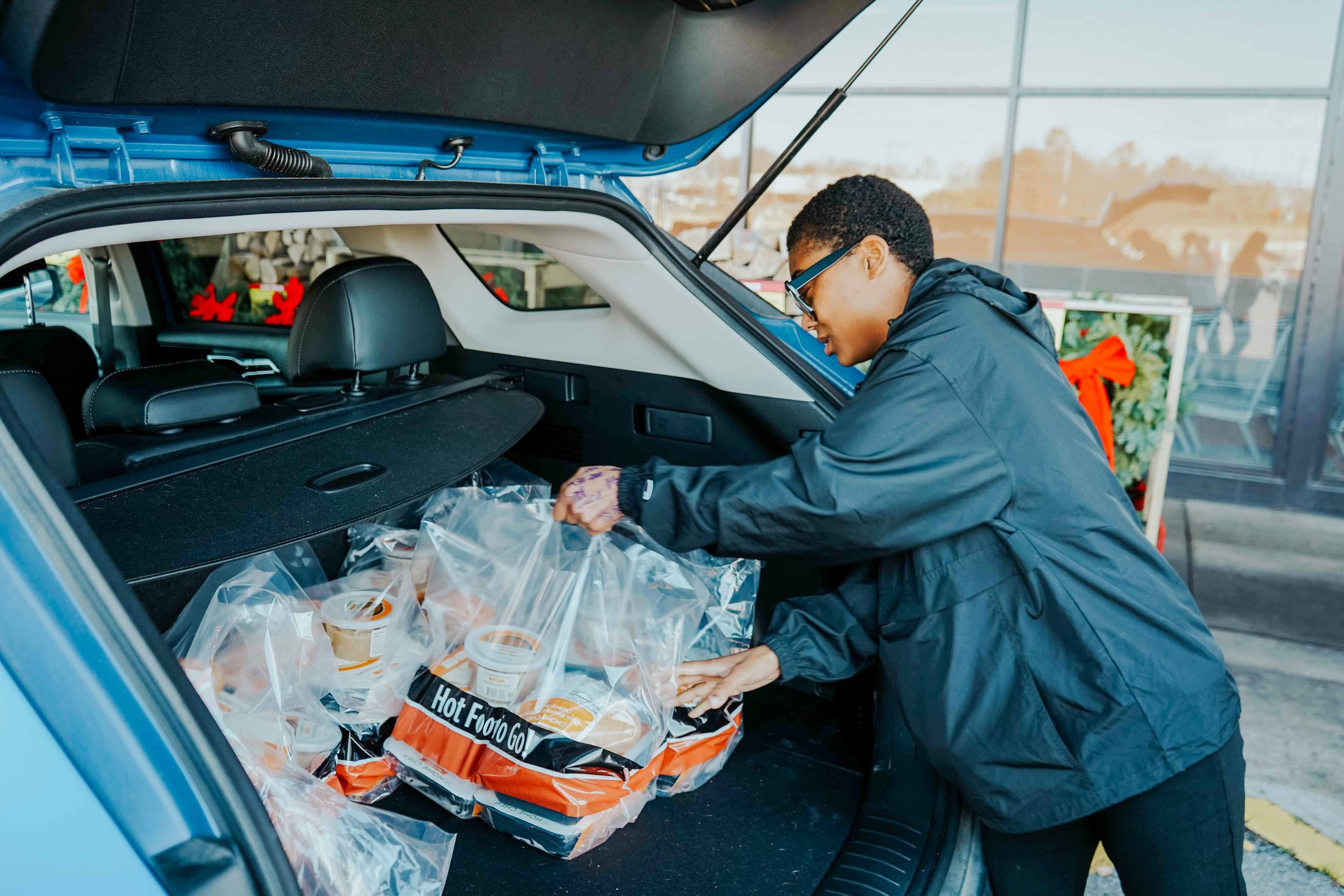 A volunteer loading bags of hot meals labeled “Hot Food to Go!” into the trunk of a car at a holiday meal drive.